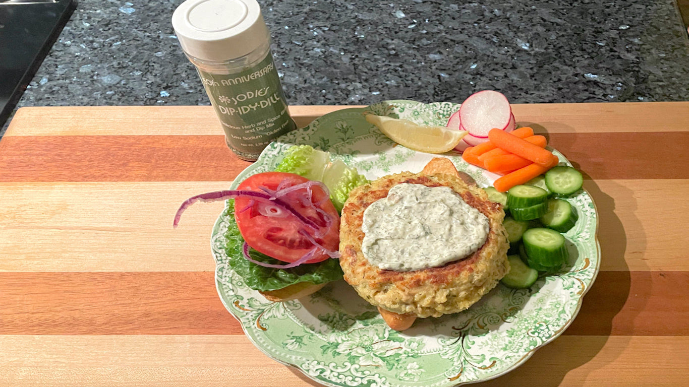 Salmon Burger made with Dip-idy Dill herb and spice blend plated with a burger roll lettuce, tomato and purple onion with fresh carrots, cucumbers, radishes and a lemon wedge.