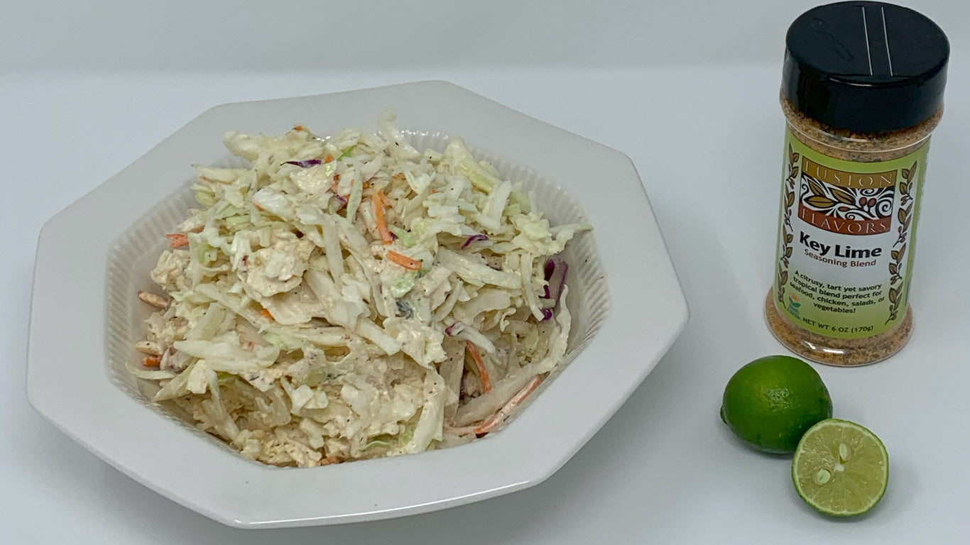 Bowl of coleslaw on white plate with a bottle of Fusion Flavors Key Lime seasoning blend and a key lime sliced in hallf.